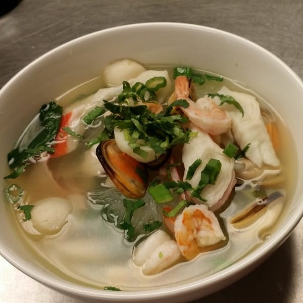 A white bowl of Thai seafood soup with shrimp, mussels, fish balls, and herbs.