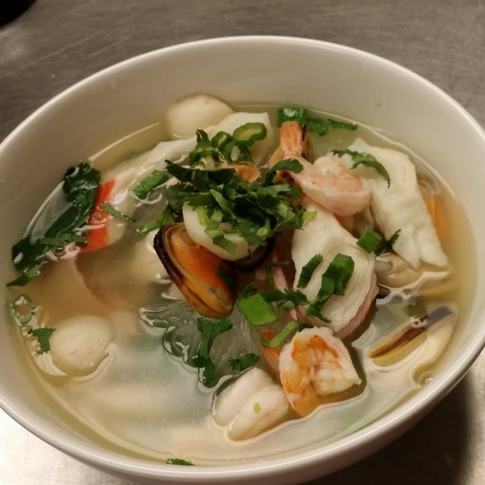 A white bowl of Thai seafood soup with shrimp, mussels, fish balls, and herbs.