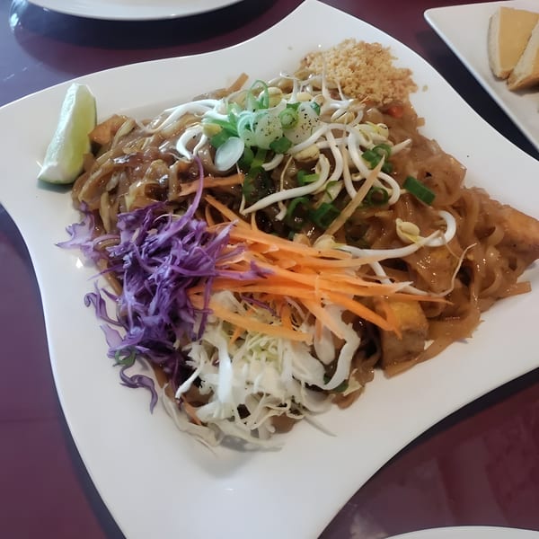 A plate of Thai Pad See Ew noodles with tofu, bean sprouts, cabbage, carrots, and a lime wedge.