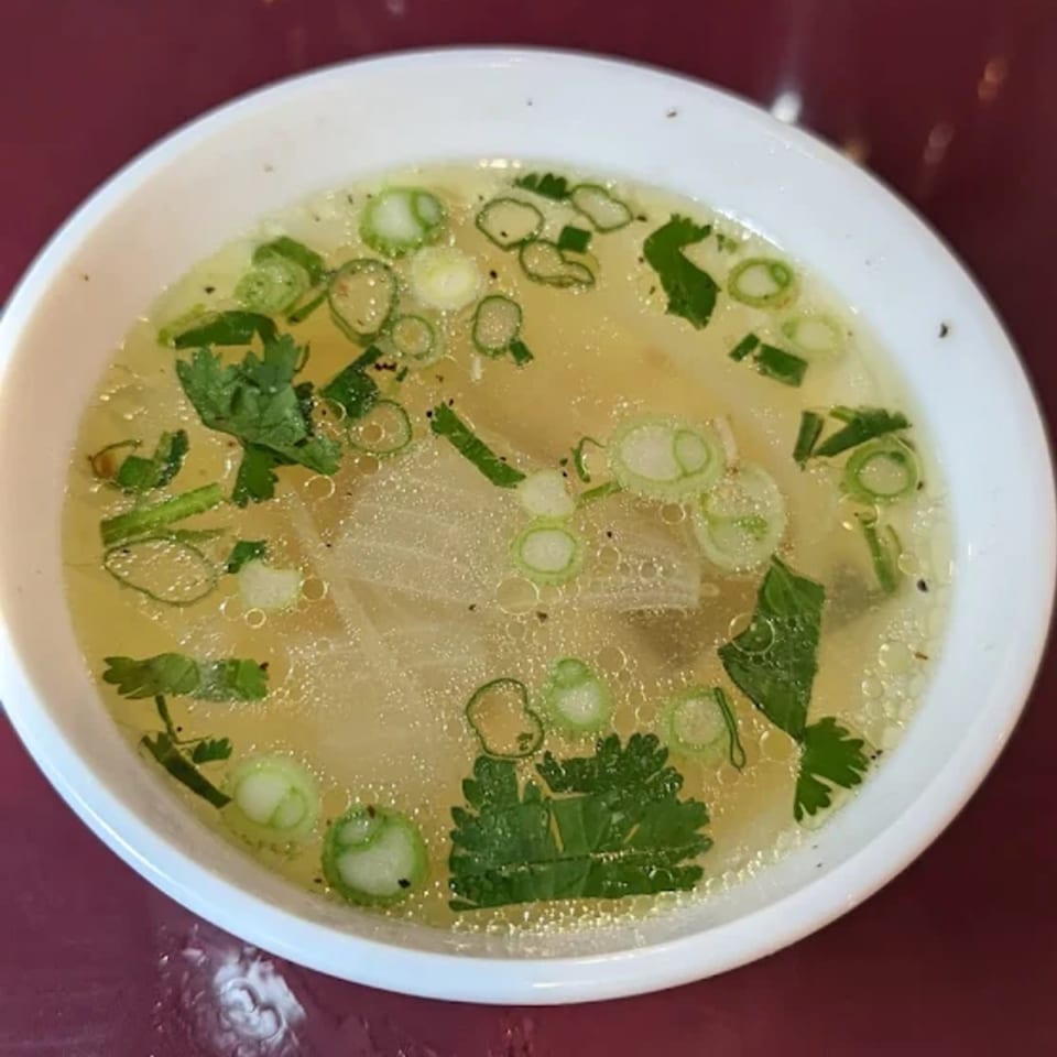 A white bowl of clear Thai soup with green onions and cilantro on a red table.